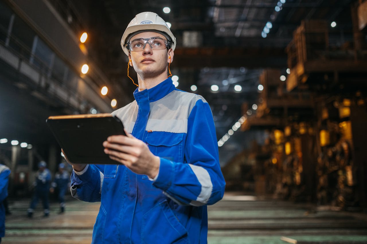 why-choose-us Engineer in safety gear uses a tablet in a large industrial factory setting, focused and diligent.